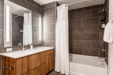 A modern bathroom features a wooden vanity with a white sink in the foreground and a white bathtub with a shower curtain against dark grey tiled walls in the background.