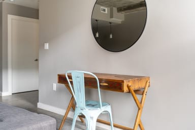 A light blue metal chair sits before a rustic wooden desk against a grey wall with a round mirror hanging above.