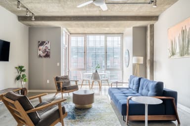 A modern living room features a blue sofa and leather armchairs in the foreground, with a dining set and large windows overlooking a city in the background.