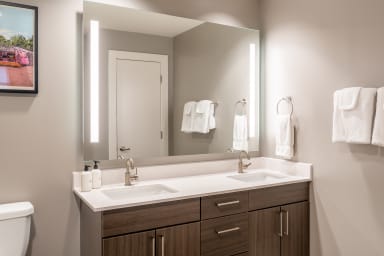 A double-sink vanity with a large illuminated mirror sits in the foreground, while a white door and hanging towels are visible in the background.