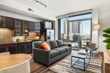 A modern open-concept living space features a grey sofa and coffee table in the foreground, with a dark wood kitchen and a city skyline visible through large glass doors in the background.