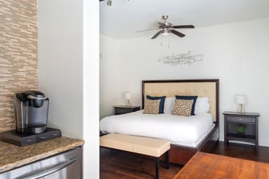 A coffee maker sits on a granite counter in the foreground, while a modern bedroom with a large bed and ceiling fan fills the background.