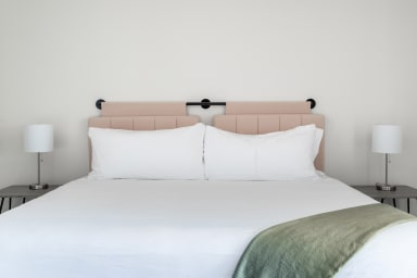 A white bed with a green throw blanket and two white pillows sits before a blush-colored headboard against a plain white wall.