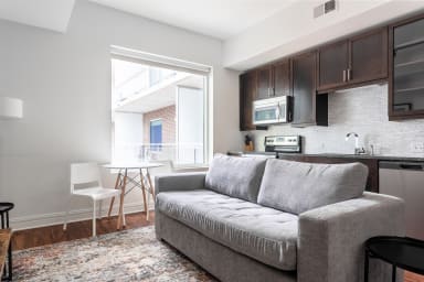 A grey sofa and patterned rug sit in the foreground of a modern studio apartment featuring a small dining set and a kitchenette in the background.