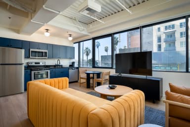 A yellow ribbed sofa and round coffee table sit in the foreground of a modern open-concept living space with a kitchen and large windows in the background.