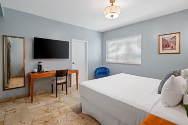 A white bed occupies the foreground of a light blue bedroom featuring a wooden desk with a TV and a mirror against the back wall.