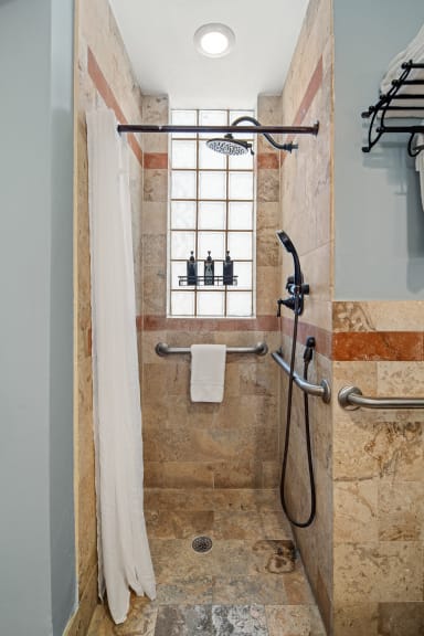 A walk-in shower with tan stone tiles and grab bars features a white curtain in the foreground and a glass block window in the background.