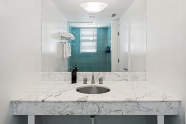 A white marble countertop with a built-in sink sits in the foreground, while a large mirror reflects a bathroom with blue tiles and a window in the background.