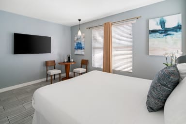 A white bed occupies the foreground of a modern bedroom featuring a small dining set, a wall-mounted TV, and a window with beige curtains in the background.