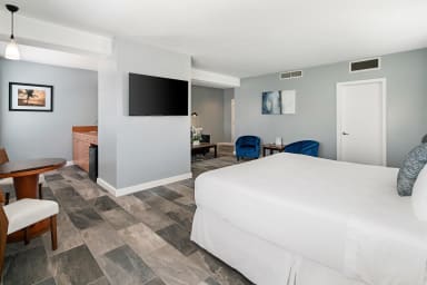 A white bed occupies the foreground of a modern hotel room featuring a wall-mounted television and a seating area with blue chairs in the background.