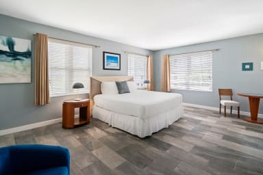 A blue armchair sits in the foreground of a bright bedroom featuring a white bed, wooden furniture, and light blue walls with shuttered windows.