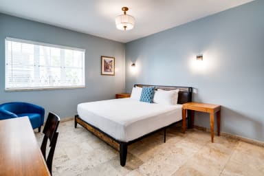 A wooden desk and chair sit in the foreground of a bedroom featuring a white-sheeted bed against light blue walls.