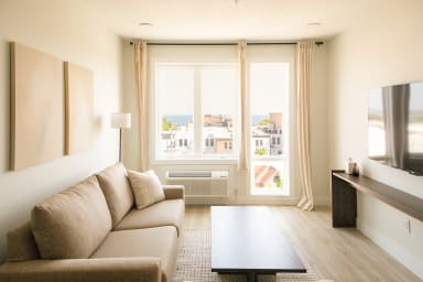 A beige sofa and dark coffee table sit in a bright living room, with a large window in the background overlooking a city skyline and the ocean.