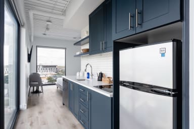 A stainless steel refrigerator and dark blue cabinetry occupy the foreground of a modern studio apartment with a living area and large window in the background.
