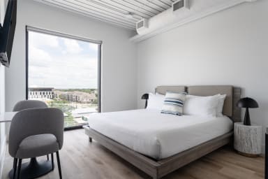 A modern bedroom features a white bed and grey chairs in the foreground, with a large window overlooking a city landscape in the background.