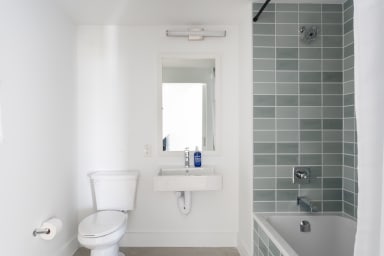 A white toilet and sink stand against a plain white wall in the foreground, while a bathtub with sage green tiles is positioned in the background.