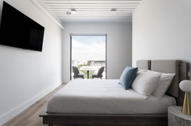 A white bed with blue and white pillows sits in the foreground of a bright room, with a television on the wall and a window overlooking a city view in the background.