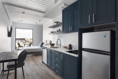 A modern studio apartment features a navy blue kitchenette in the foreground and a bright living area with a bed and window in the background.