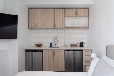 A white bed is in the foreground of a small kitchenette featuring light wood cabinets, a white tiled backsplash, and stainless steel appliances.