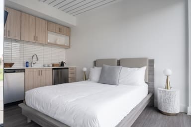 A white bed with grey pillows sits in the foreground of a modern studio apartment featuring a small kitchenette in the background.