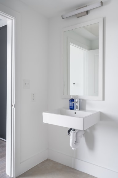 A white wall-mounted sink with a soap dispenser sits in the foreground, while a mirror and light fixture are mounted on the white wall in the background.