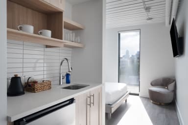 A small kitchenette with a sink and open shelving sits in the foreground, leading into a bright studio room with a bed, a grey armchair, and a large window in the background.