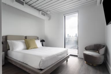 A modern bedroom features a white bed with a gold accent pillow in the foreground and a grey armchair beside a tall window overlooking a city skyline in the background.