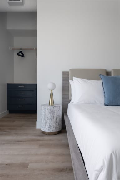 A white bed with a blue pillow sits beside a marble side table and lamp, with a walk-in closet featuring a dark blue dresser in the background.