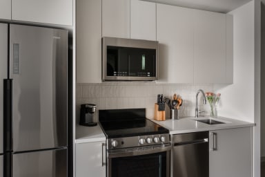 A stainless steel refrigerator and oven stand in the foreground of a modern kitchen with white cabinetry and a tiled backsplash in the background.