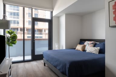 A modern bedroom features a bed with a blue quilt and decorative pillows in the foreground, with a glass door leading to a balcony and city buildings in the background.