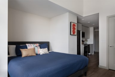 A bed with a blue quilted comforter and decorative pillows sits in the foreground, while a small kitchen area is visible in the background.