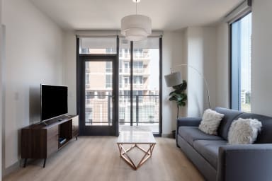 A modern living room features a grey sofa, white coffee table, and TV stand in the foreground, with a glass door leading to a balcony overlooking city buildings in the background.