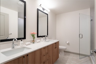 A modern bathroom features a double vanity with wooden cabinets and white countertops in the foreground, with a toilet and a glass shower enclosure in the background.