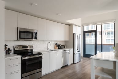 A modern white kitchen with stainless steel appliances occupies the foreground, leading toward a glass door and balcony in the background.