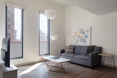 A modern living room features a grey sofa and a geometric marble coffee table in the foreground, with white walls, a piece of abstract art, and large windows overlooking city buildings in the background.