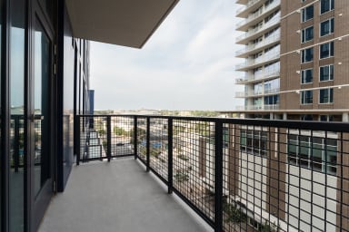 A concrete balcony with a black metal railing overlooks a city skyline and a tall brick apartment building.