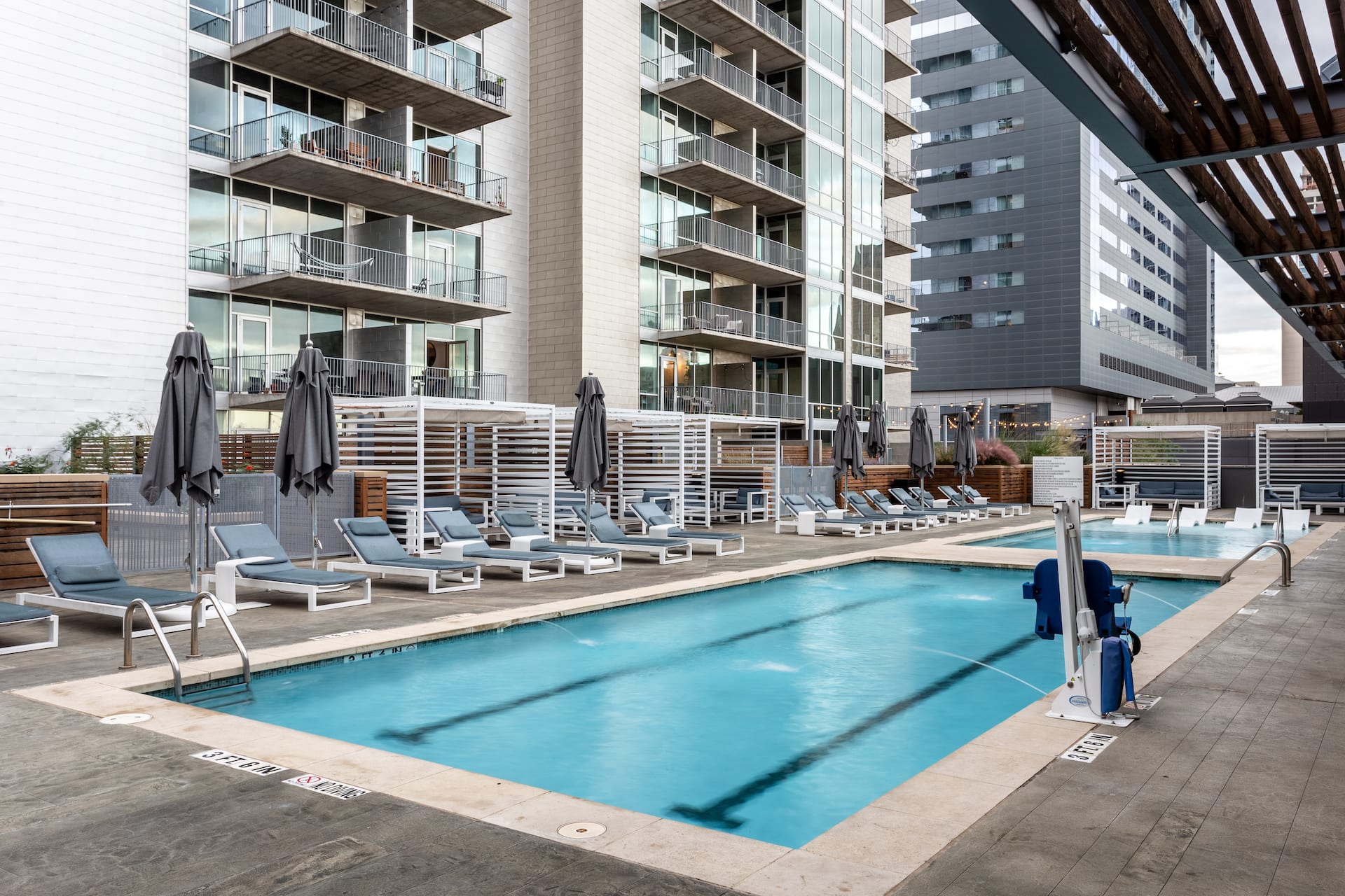 A swimming pool with lounge chairs and umbrellas sits in the foreground, set against a backdrop of modern high-rise apartment buildings.