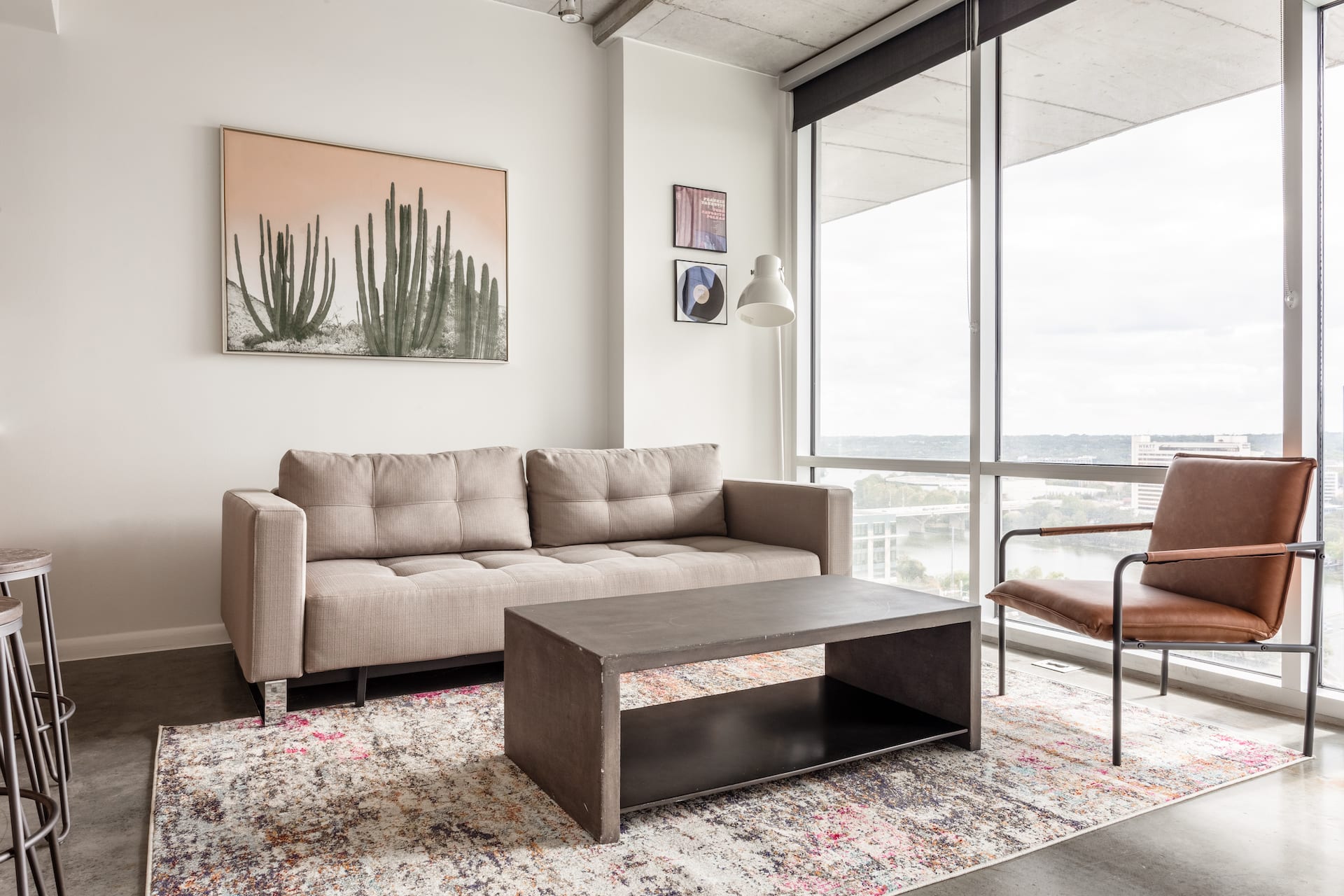A modern living room features a beige sofa, a dark coffee table, and a brown leather chair on a patterned rug, with a large window overlooking a city skyline in the background.