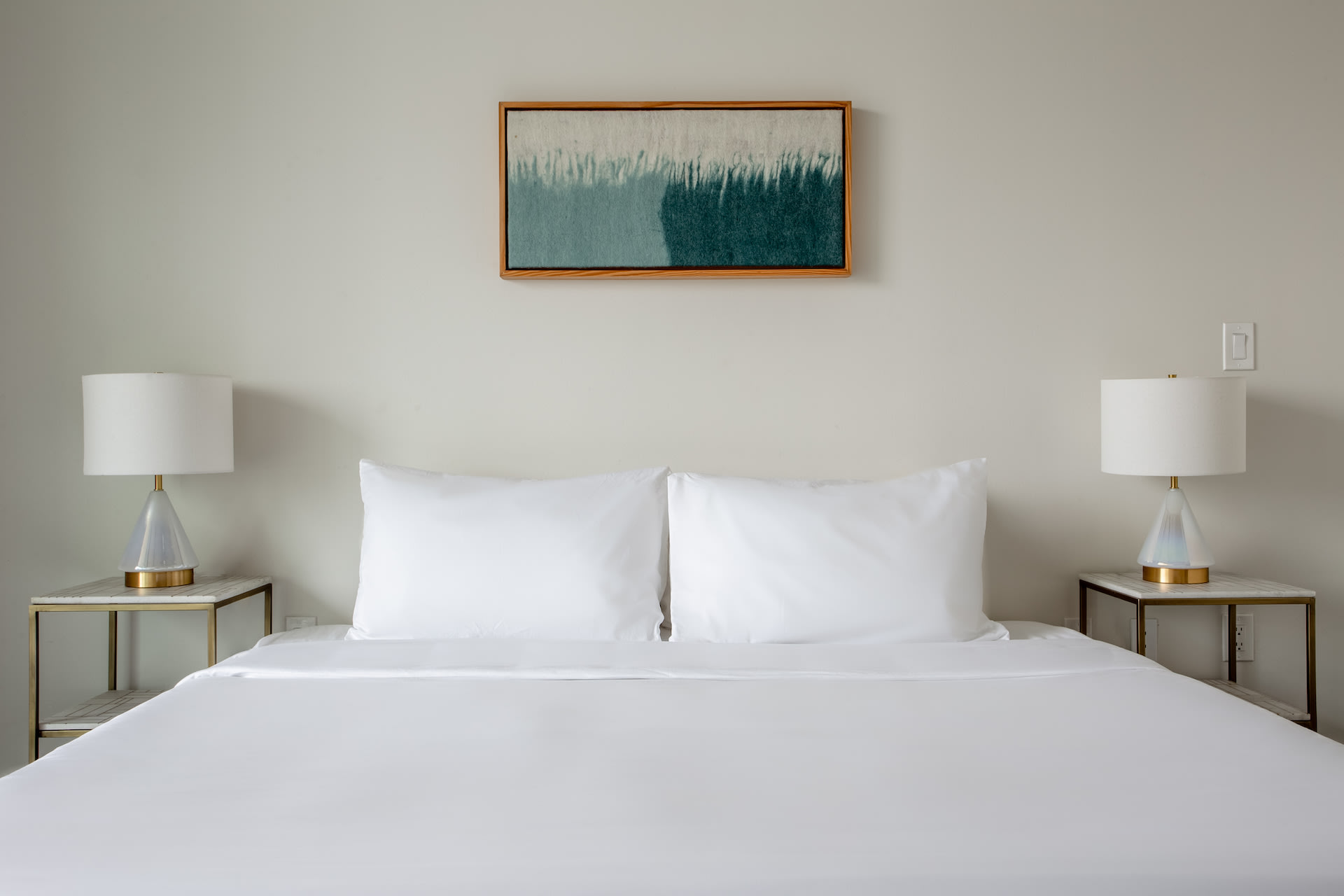 A white bed with two pillows sits in the foreground, flanked by matching bedside tables and lamps, while a framed abstract painting hangs on the neutral wall in the background.