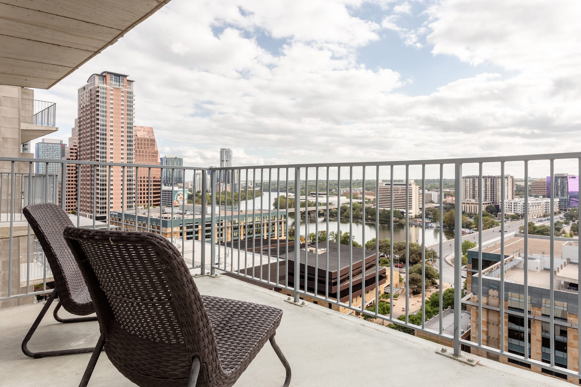 Two brown wicker chairs sit on a concrete balcony overlooking a city skyline and a river under a cloudy sky.