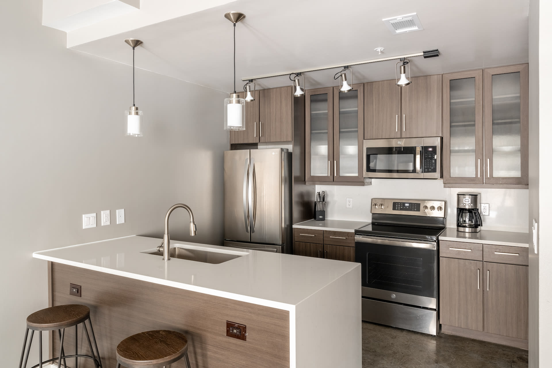 A modern kitchen features a white quartz island with wooden stools in the foreground and stainless steel appliances with grey cabinetry in the background.
