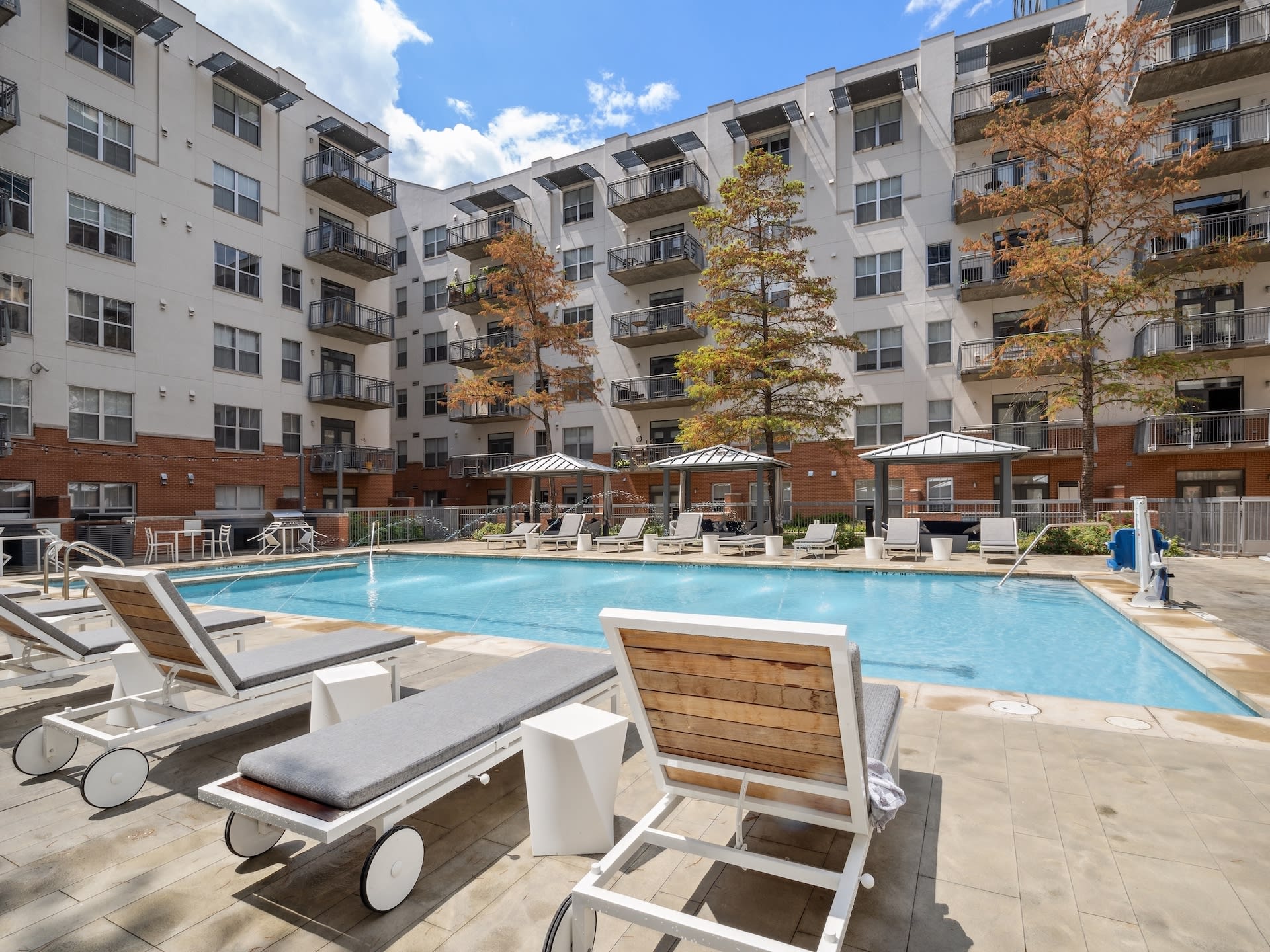 White lounge chairs line the edge of a bright blue swimming pool, surrounded by modern apartment buildings and autumn trees under a sunny sky.