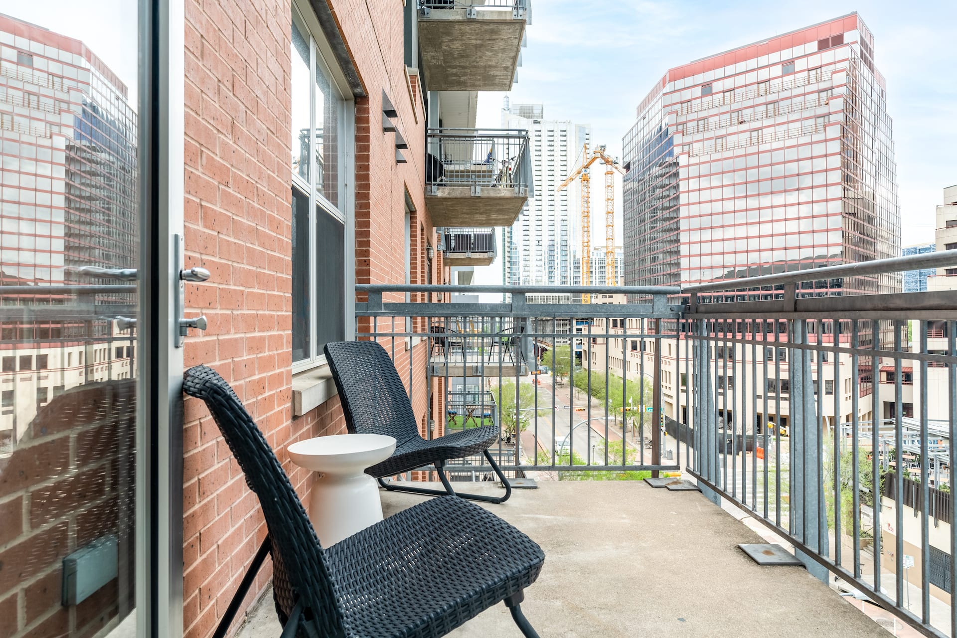 Two black wicker chairs and a small white table sit on a concrete balcony against a brick wall, overlooking a city skyline with tall buildings and a construction crane.