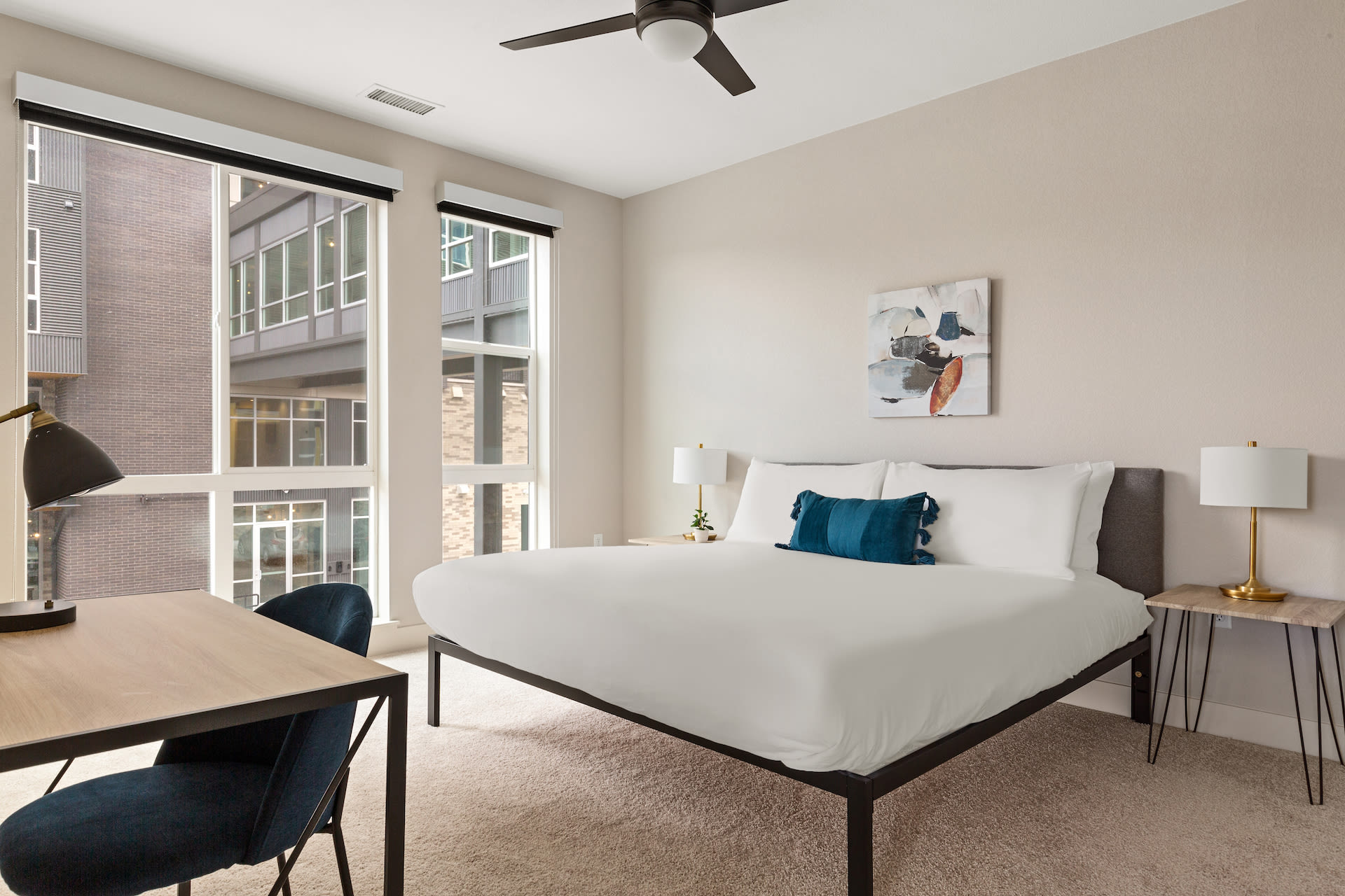A modern bedroom features a white-linened bed with a blue accent pillow in the foreground, set against a neutral-toned wall with a piece of abstract art and large windows overlooking city buildings in the background.