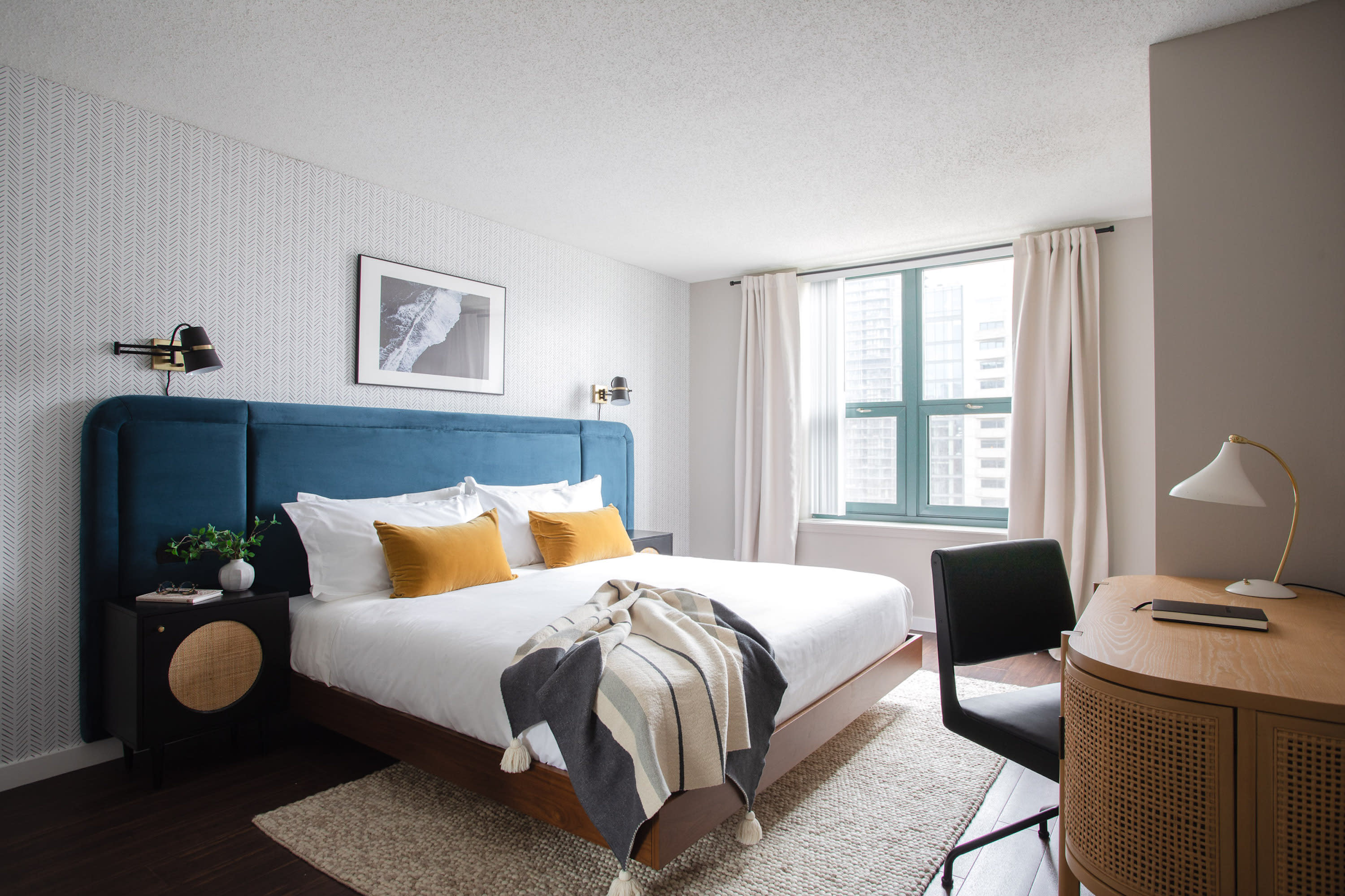 A modern bedroom features a bed with a blue velvet headboard and yellow pillows in the foreground, set against a background of patterned wallpaper and a window with white curtains.