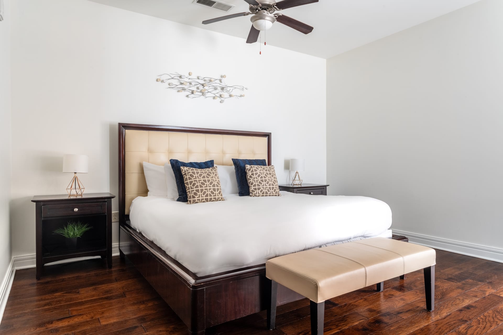 A large bed with white linens and decorative pillows sits in the center of a room with dark wood floors, flanked by nightstands and a beige bench in the foreground against a plain white wall and ceiling fan in the background.