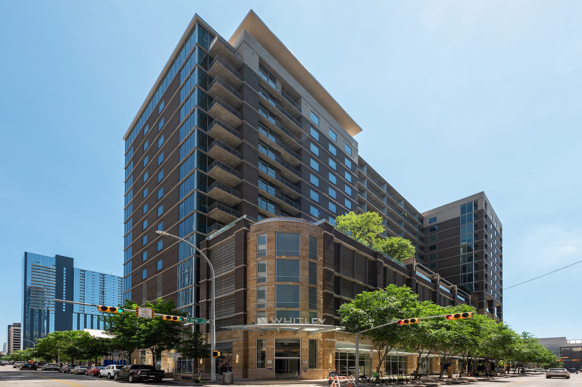 A modern high-rise residential building stands prominently along a city street lined with green trees and parked cars under a clear blue sky.