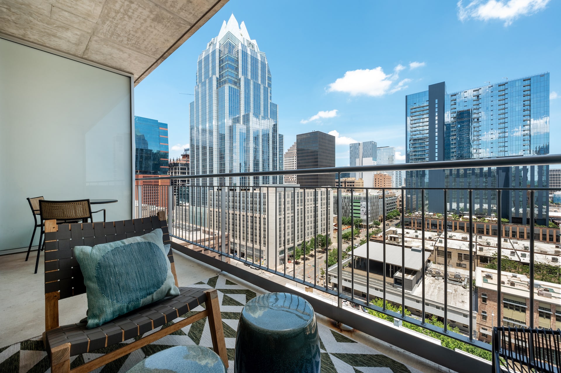 A modern balcony with a lounge chair and stool overlooks a dense city skyline featuring tall skyscrapers under a blue sky.