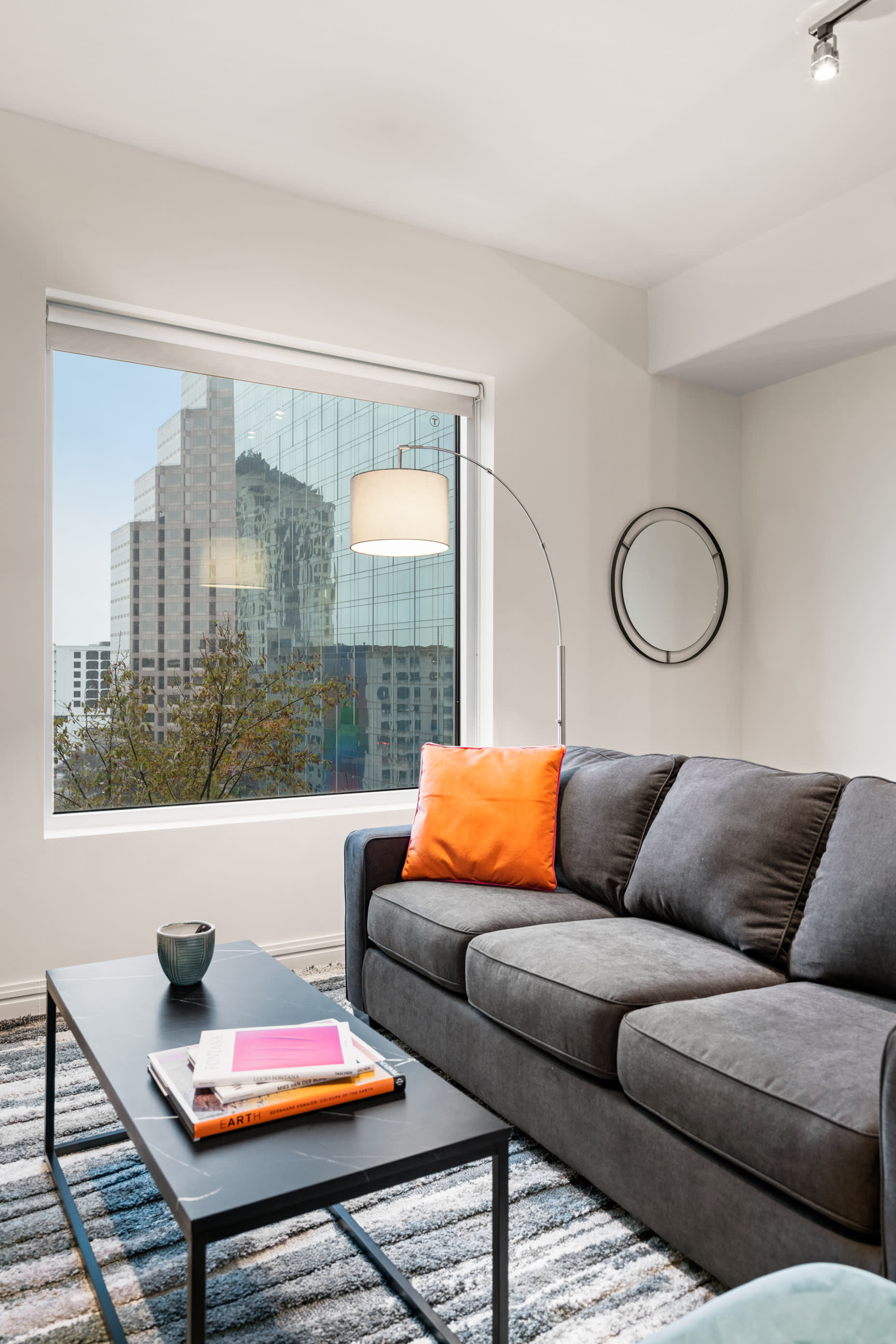 A grey sofa with an orange pillow and a black coffee table sit in the foreground of a bright room, with a large window overlooking city skyscrapers in the background.