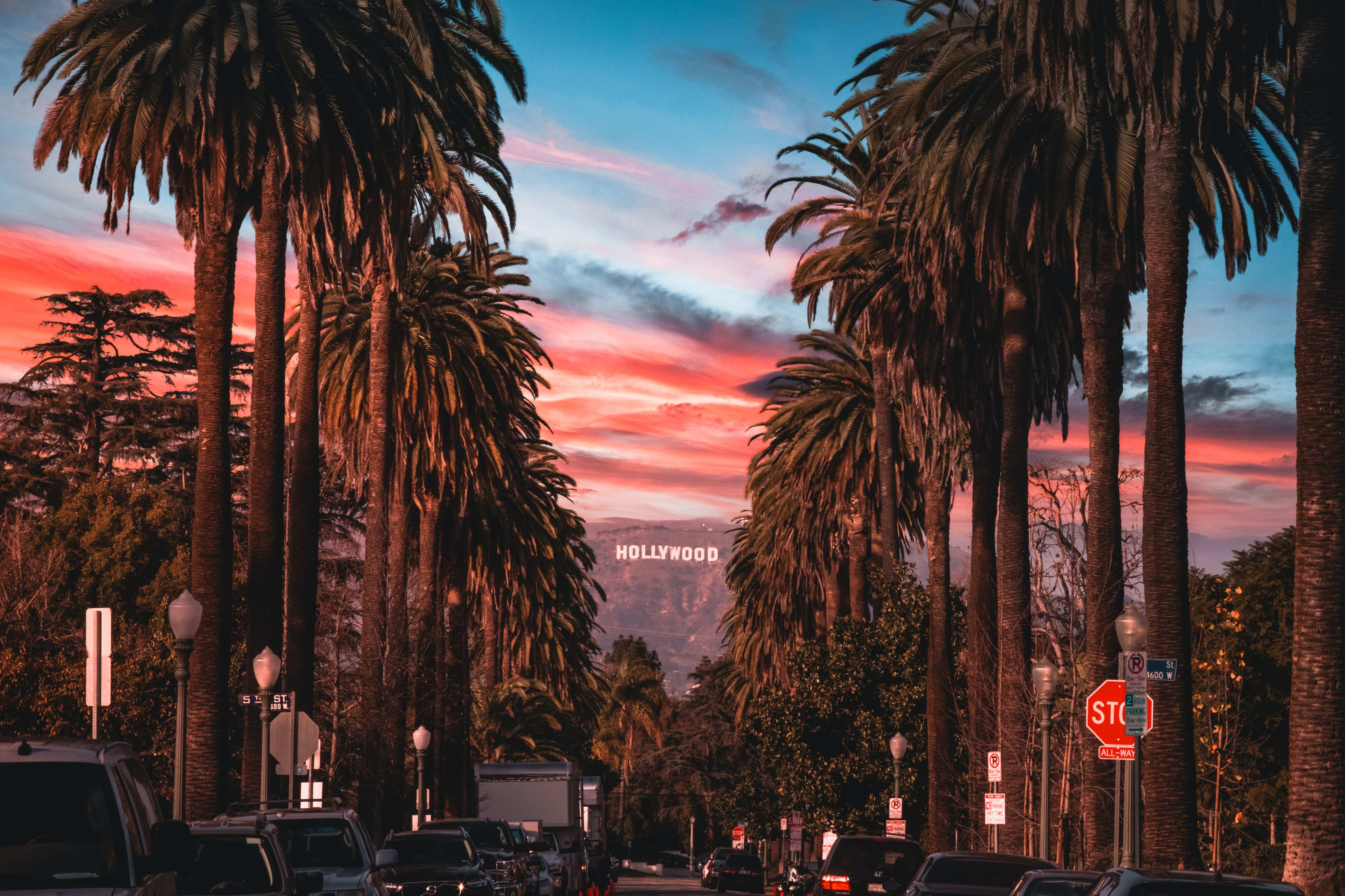 Tall palm trees line a street with parked cars in the foreground, while the Hollywood sign sits atop a distant hill under a vibrant red and blue sunset sky.